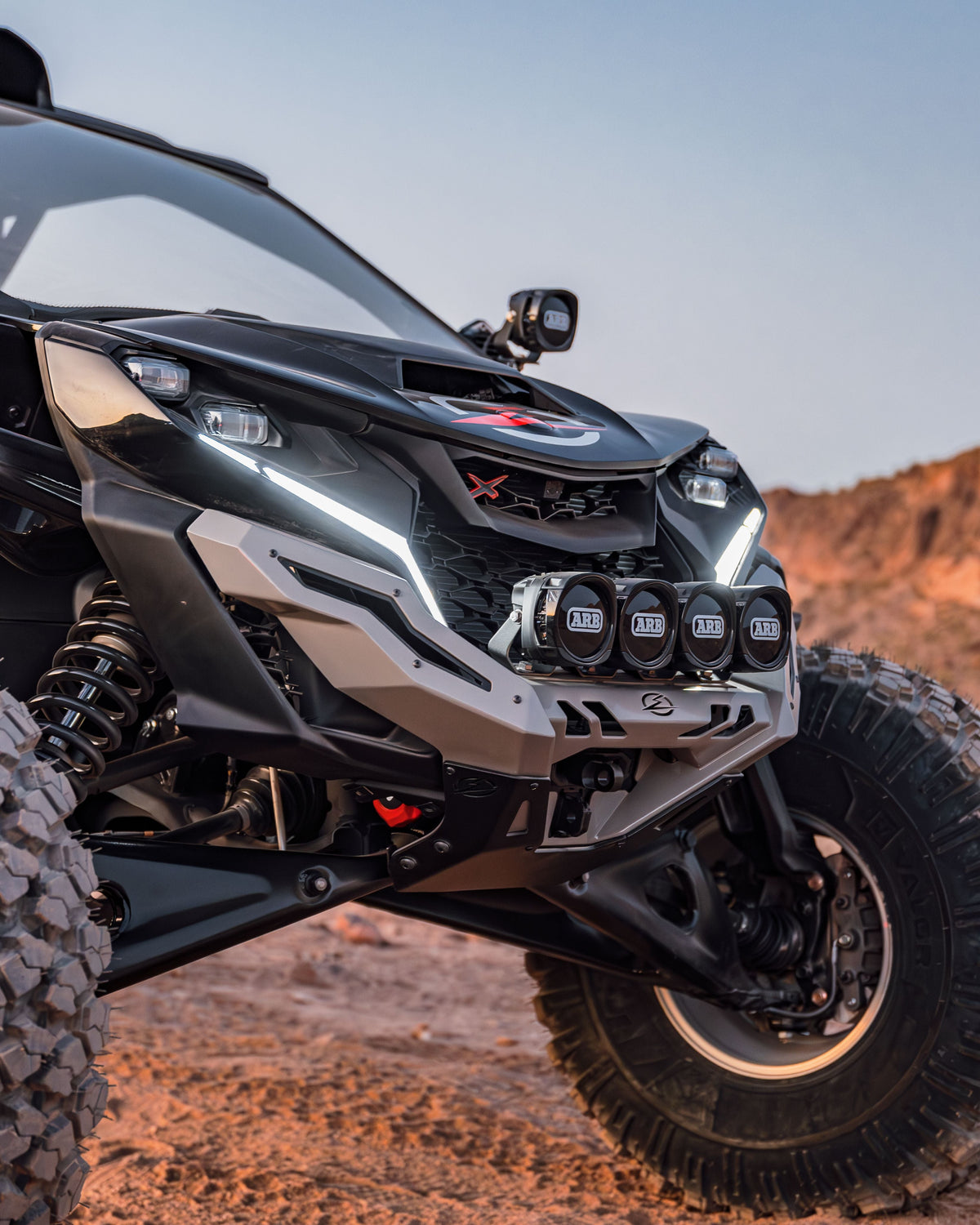 A front winch bumper designed for a Can Am Maverick R, featuring a black powder coat finish and integrated LED light bar, mounted on a off-road vehicle.