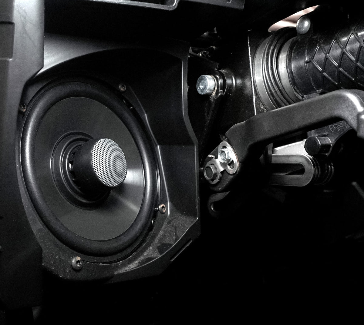 Close up of a circular vehicle speaker mounted in a black plastic housing next to metal mechanical components